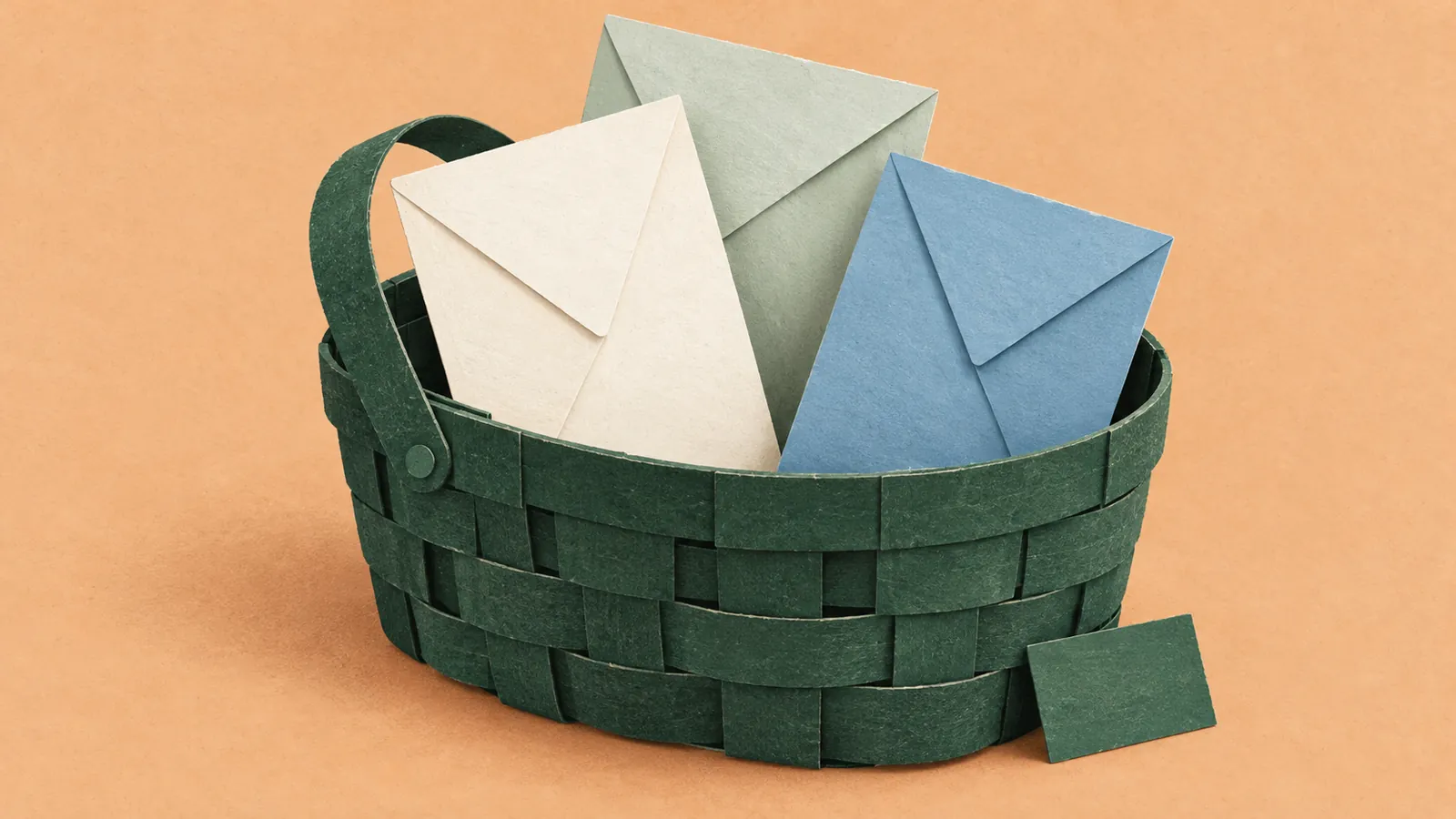 A forest-green woven paper basket holds blank ivory, sage, and blue envelopes on an apricot paper background.